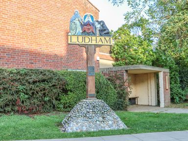 A wooden Ludham village sign on a stone base near a bus shelter and bushes at 27 Whitegates in Ludham