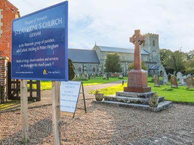 A churchyard with a stone church tower a large cross monument and informational signs at St Catherines Church in Ludham