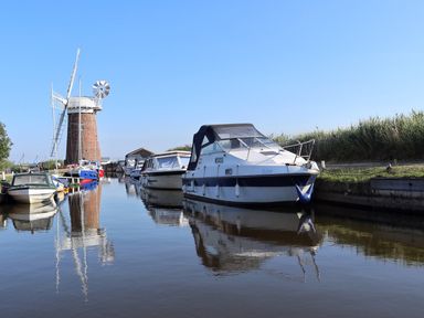 A canal with boats docked along the banks and a windmill in the background at 27 Whitegates in Ludham