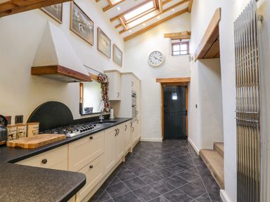 A narrow kitchen with white cabinets black countertops a gas stove a sink a black door and framed pictures on the wall at Pen Y Bryn Cottage in Llandudno