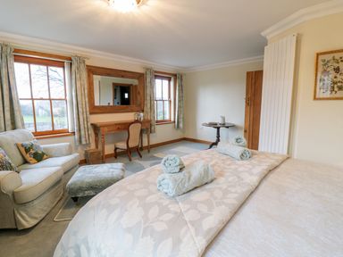 A bedroom with a bed with rolled towels a sofa an ottoman a dressing table with a chair and two windows at Pen Y Bryn Cottage in Llandudno