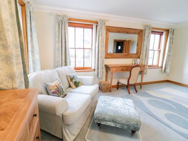 A sitting room with a sofa, ottoman, wooden desk, chair, mirror, and windows with curtains at Pen Y Bryn Cottage in Llandudno