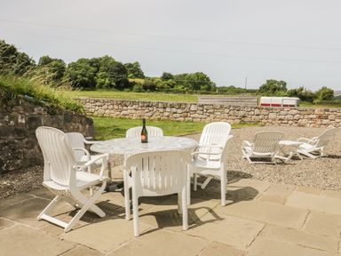 An outdoor patio with white plastic chairs around a round table with a bottle on top and lounge chairs in a gravel area with a stone wall and grassy fields beyond at Pen Y Bryn Cottage in Llandudno
