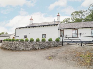 A white cottage with two chimneys a stone wall green bushes and a black metal gate at Pen Y Bryn Cottage in Llandudno