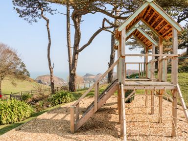 A wooden play structure with climbing ropes and a slide in a park with trees and hills in the background at Martin Cottage in Ilfracombe