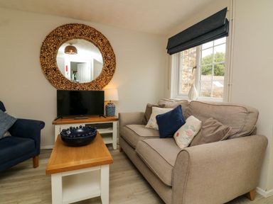 A living room with a beige sofa and cushions a blue armchair a wooden coffee table a TV on a stand and a round decorative mirror on the wall at Martin Cottage in Ilfracombe