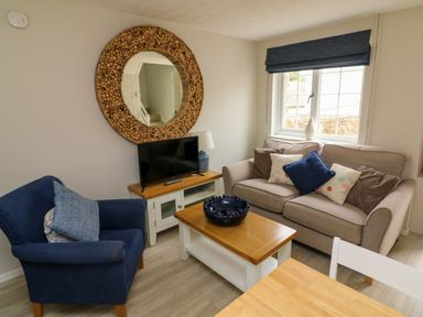 A living room with a beige sofa with cushions a blue armchair a wooden coffee table a television on a wooden TV stand and a round wooden framed mirror on the wall at Martin Cottage in Ilfracombe