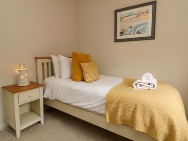 A single bed with white sheets, yellow pillows and a yellow blanket with towels on top next to a nightstand with a lighthouse lamp and a framed picture on the wall at Martin Cottage in Ilfracombe