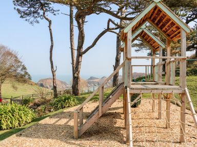 A wooden play structure with a slide and climbing ropes in a garden with trees and ocean view at Swallow Cottage in Ilfracombe