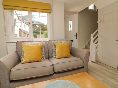 A living room with a beige sofa with yellow cushions near a window and a white door at Swallow Cottage in Ilfracombe