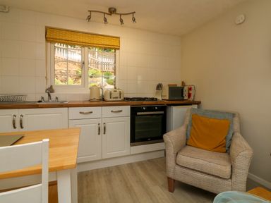 A kitchen with white cabinets a wooden countertop window with yellow blind and an armchair with cushions at Swallow Cottage in Ilfracombe