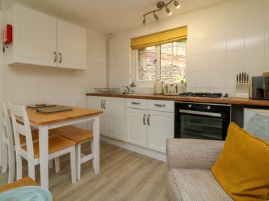 A kitchen with white cabinets wooden countertops a dining table with two chairs and a beige sofa with a yellow cushion at Swallow Cottage Ilfracombe