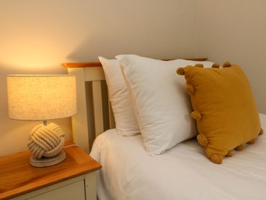 A bed with white pillows and a yellow cushion next to a wooden bedside table with a lamp at Swallow Cottage in Ilfracombe