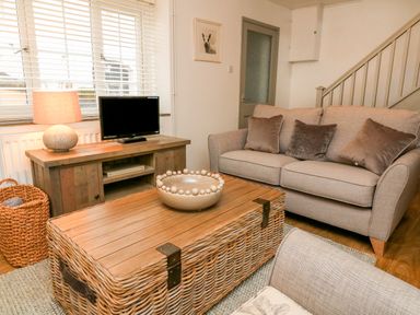 A living room with a beige sofa and cushions a wooden TV stand with a lamp a wicker basket and a woven coffee table with a decorative bowl at Kingfisher Cottage in Ilfracombe