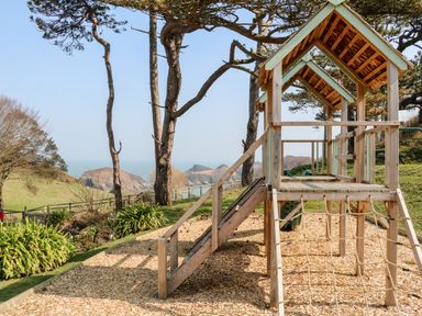 A wooden playground structure with a slide and climbing net in a garden with trees and a view of the sea at Kingfisher Cottage in Ilfracombe