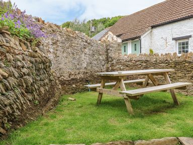 A wooden picnic table on grass beside stone walls with a white building in the background at Partridge Cottage in Ilfracombe
