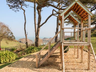A wooden playground structure with ropes and a slide in a garden with trees and hills in the background at Partridge Cottage in Ilfracombe