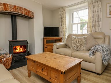 A living room with a wood burning stove a wooden coffee table a beige leather sofa and a television on a wooden stand at Partridge Cottage in Ilfracombe
