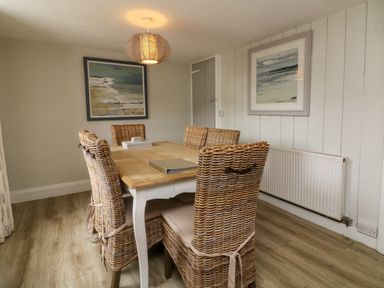A dining room with a wooden table and six wicker chairs and paintings on the wall at Partridge Cottage in Ilfracombe