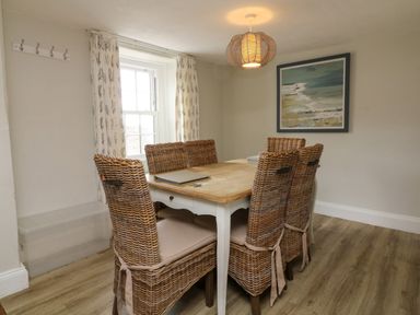 A dining room with a wooden table and six wicker chairs near a window with curtains at Partridge Cottage in Ilfracombe