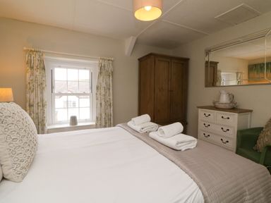 A bedroom with a bed with white linens and folded towels a wooden wardrobe a chest of drawers and a window with floral curtains at Partridge Cottage in Ilfracombe