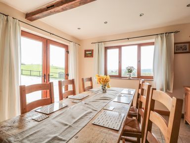 A dining room with a wooden table and chairs flowers in vases on the table and windows at Blaen Henllan in Builth Wells