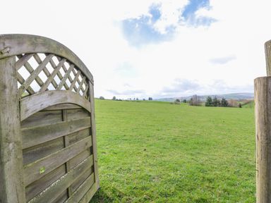 A wooden gate opening to a grassy field with trees and hills in the distance at Blaen Henllan in Builth Wells