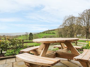 A wooden round picnic table with benches on a stone patio overlooking green fields and trees at Blaen Henllan in Builth Wells