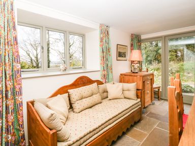 A room with a wooden daybed with cushions beside windows with floral curtains and a wooden cabinet with a lamp at Timber Barn in Eardisland