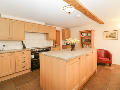 A kitchen with wooden cabinets a kitchen island with flowers a black stove a wooden bookshelf and a red chair at Timber Barn in Eardisland