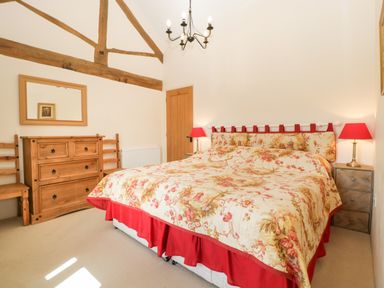 A bedroom with a bed with floral bedding two red lamps a wooden dresser two chairs and exposed timber beams at Timber Barn in Eardisland