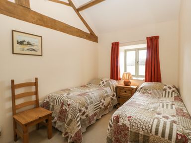A bedroom with two single beds a wooden chair a nightstand with a lamp and a window with red curtains at Timber Barn in Eardisland