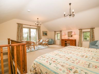 A bedroom with a patterned bedspread a wooden desk with lamps a seating area with wicker chairs and a table and windows with curtains at Timber Barn in Eardisland