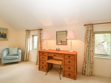 A room with a wicker desk with two lamps and a stool between two windows with curtains and a blue armchair at Timber Barn in Eardisland