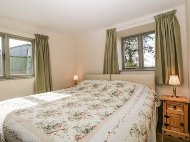A bedroom with a bed covered by a floral quilt two windows with green curtains and two wooden bedside tables with lamps at Timber Barn in Eardisland