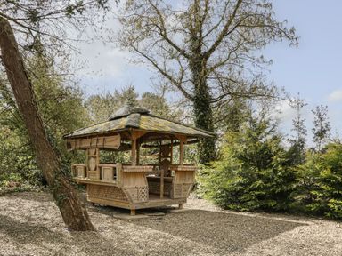 A wooden gazebo with a thatched roof in a garden area surrounded by trees and bushes at Timber Barn in Eardisland