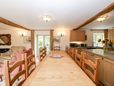 A dining area with a long wooden table and chairs next to a kitchen with wooden cabinets and sunflowers in a vase at Timber Barn in Eardisland