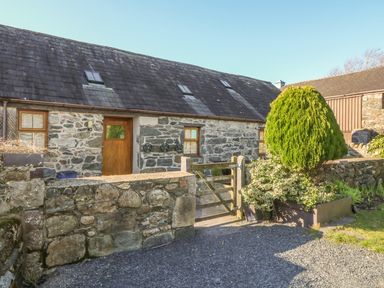 Stone cottage with wooden door and windows a gravel driveway and a wooden gate next to a planter with a shrub at Beudy Bach in Y Felinheli