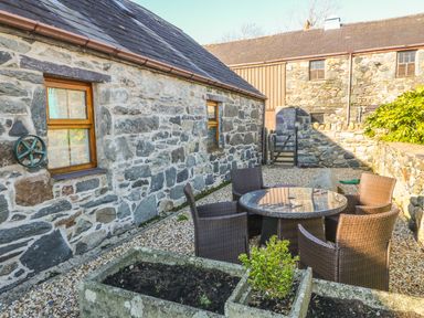 An outdoor patio area with a round glass table and four wicker chairs next to stone buildings with wooden windows at Beudy Bach in Y Felinheli