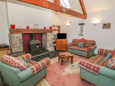 A living room with plaid armchairs and sofas around a wood stove and a television on a wooden cabinet at Beudy Bach in Y Felinheli