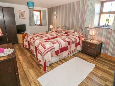 A bedroom with a double bed covered in a patchwork quilt bedside tables lamps a wardrobe television and two windows at Beudy Bach in Y Felinheli