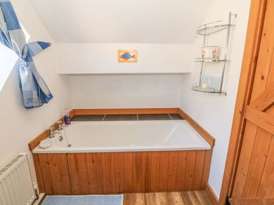 A bathroom with a bathtub with wooden paneling a small window with blue curtains and a wooden door at Beudy Bach in Y Felinheli