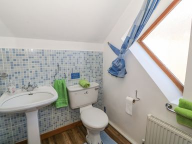 A bathroom with a pedestal sink, toilet, tiled wall, and an angled window with blue curtain at Beudy Bach in Y Felinheli