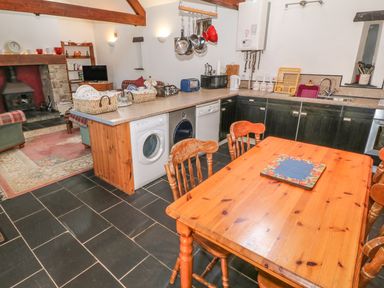 A kitchen with a wooden dining table and chairs a countertop with appliances and hanging pots in an open plan room with a fireplace visible at Beudy Bach in Y Felinheli