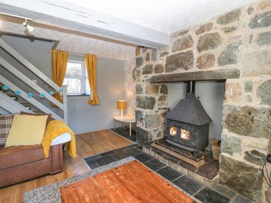 A living room with a stone fireplace and wood-burning stove a brown sofa with cushions and throw and yellow curtains at Hendre Y Felinheli