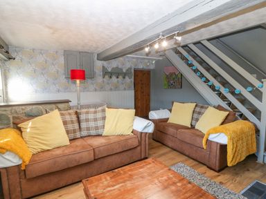 A living room with two brown sofas with cushions and throws a wooden coffee table a floor lamp with a red shade a wooden staircase with string lights and a painting of a cow on the wall at Hendre in Y Felinheli