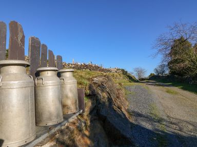 Metal milk churns next to a gravel path with stone fence and trees along the road at Hendre in Y Felinheli
