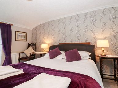 A bedroom with a double bed two purple pillows two bedside tables with lamps a wooden chair a framed picture and purple curtains at Castle Lodge in Borrowdale