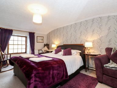 A bedroom with a double bed purple throws and cushions a window with purple curtains a chair and bedside tables with lamps at Castle Lodge in Borrowdale