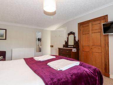 A bedroom with a bed covered by a purple blanket and white towels on top a wooden dresser with a mirror a wooden closet door and a wall-mounted television at Castle Lodge in Borrowdale